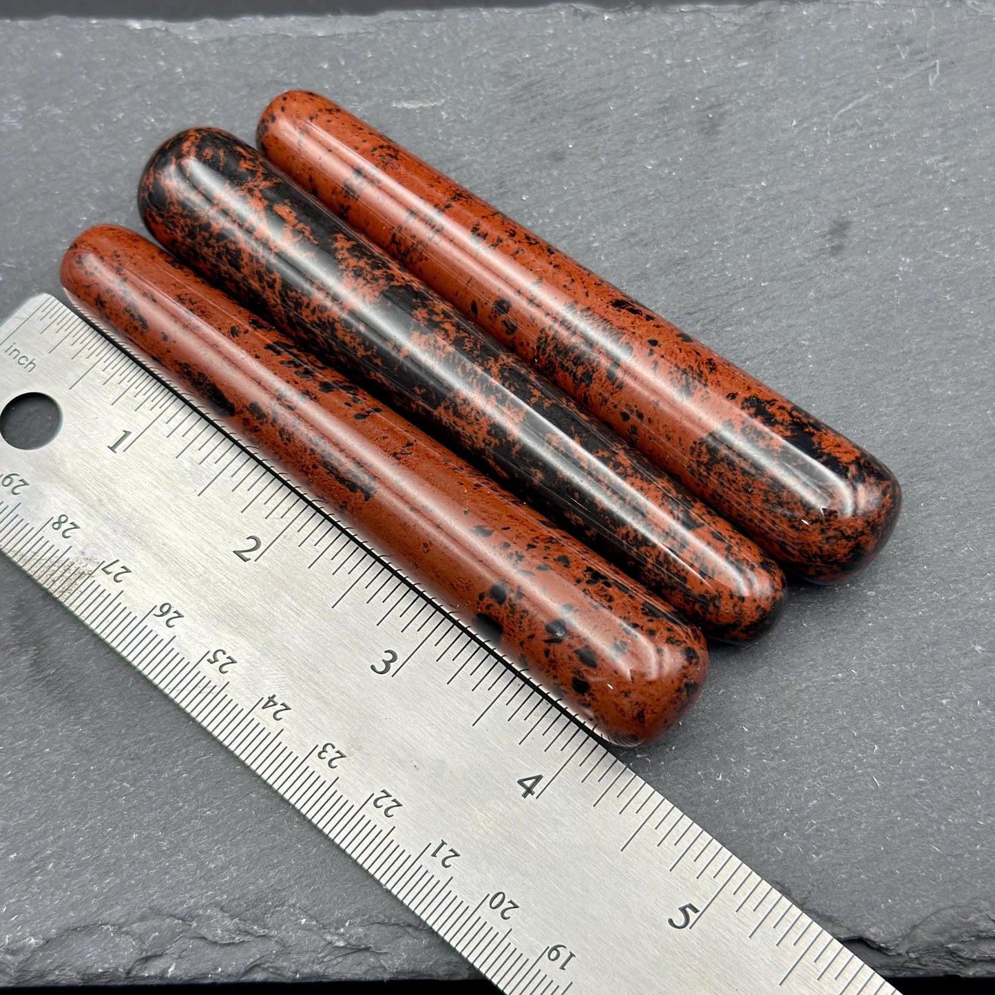 May include: Three polished red obsidian wands with black speckles. A metal ruler with inch measurements is placed next to the wands. The wands are approximately 5 inches long.