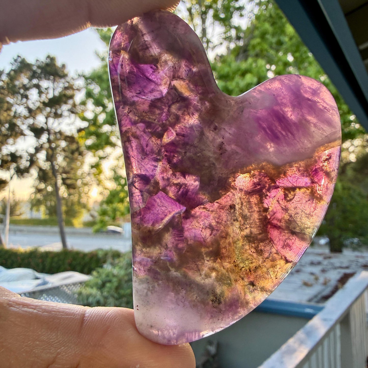 May include: A heart-shaped polished stone featuring translucent layers of deep purple, violet, and golden-brown mineral inclusions. The surface is smooth and glossy, allowing light to pass through the crystalline structure to reveal internal patterns.