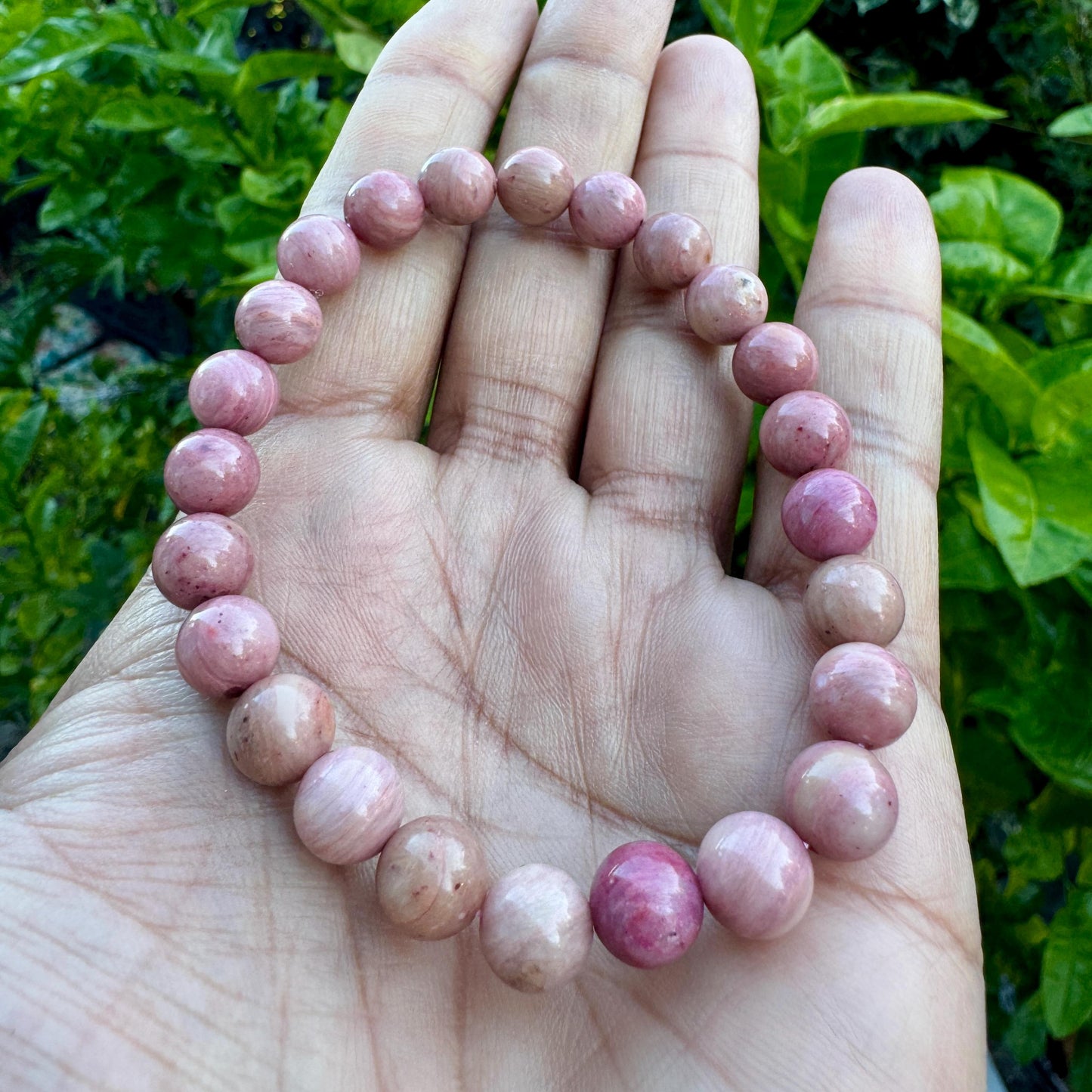 May include: A beaded bracelet composed of numerous smooth, spherical stones in varying shades of dusty rose, mauve, and soft pink. The beads are uniform in size and strung together in a circular loop, displaying natural color variations and textures.