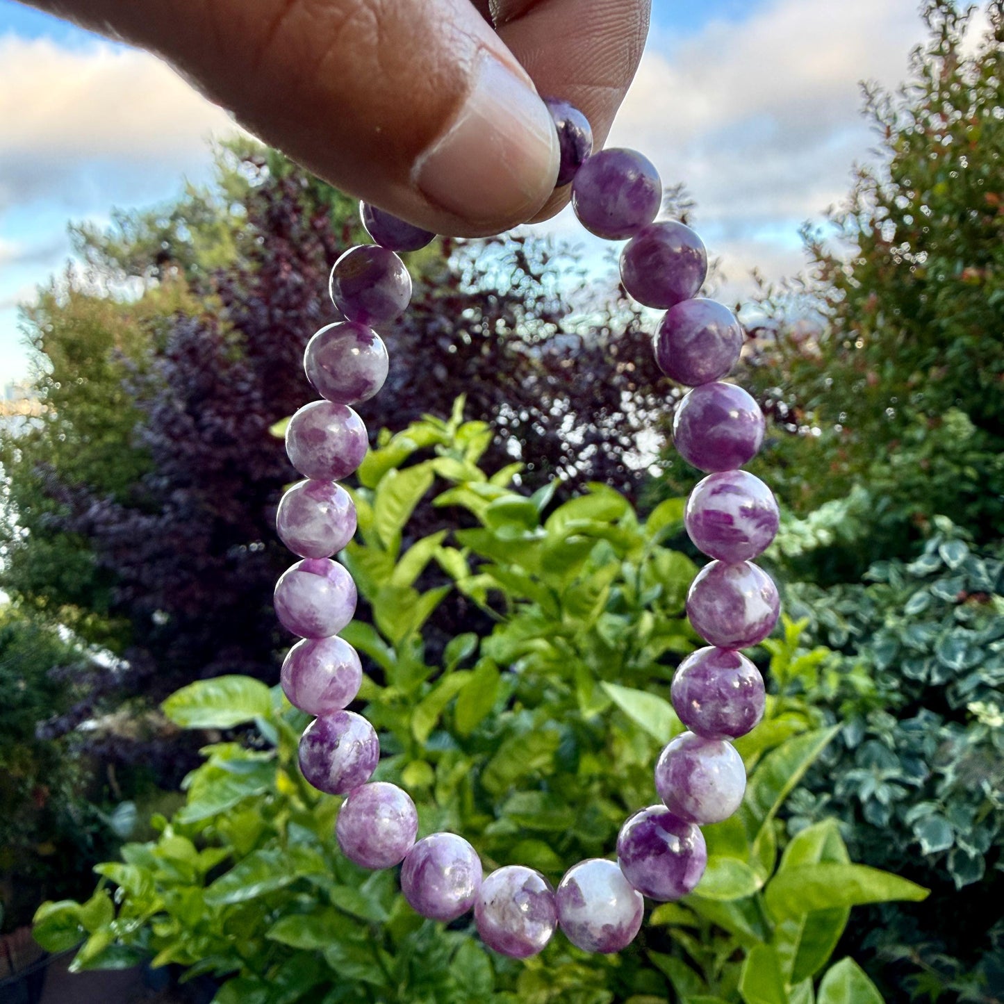 May include: A beaded bracelet composed of round, polished stones in varying shades of purple and white. The beads are strung together in a circular formation, displaying a marbled texture with translucent and opaque sections throughout the piece.