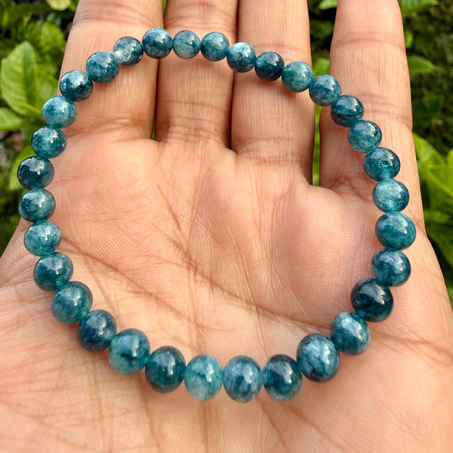 May include: A circular stretch bracelet composed of numerous small, polished, round beads in varying shades of deep teal and blue-green. The beads feature a translucent quality with subtle internal inclusions and a smooth, reflective surface finish.