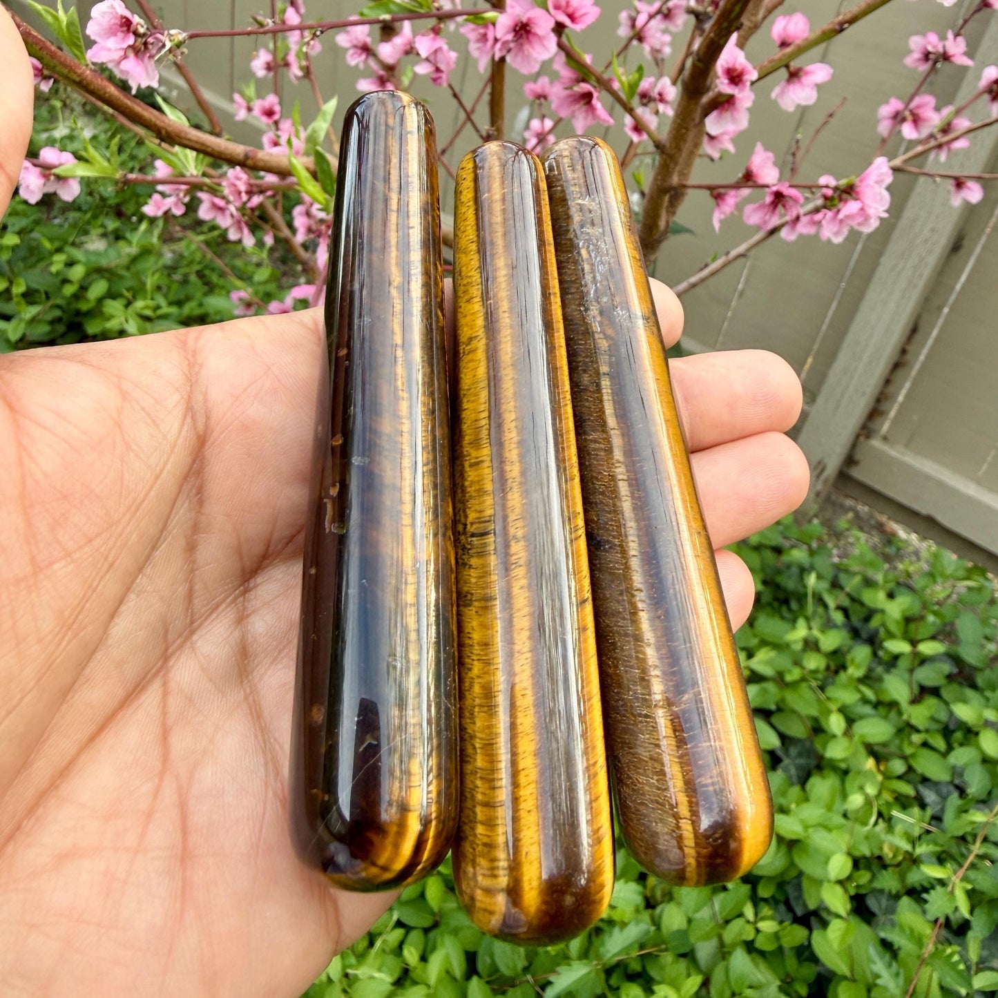 Three tiger eye stones held in a hand with a blurred background of greenery and pink flowers.