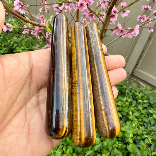 Three tiger eye stones held in a hand with a blurred background of greenery and pink flowers.