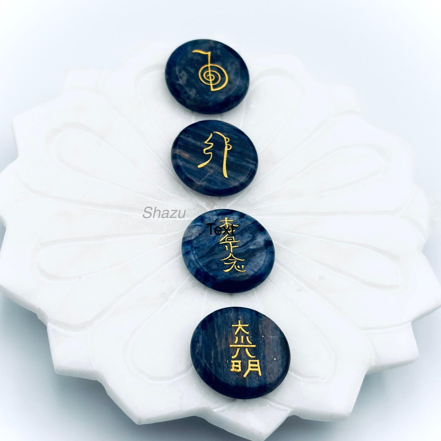 May include: Four dark blue, round stones with gold symbols and characters are arranged on a white, flower-shaped dish. The stones have a marbled appearance. The symbols and characters are in gold.