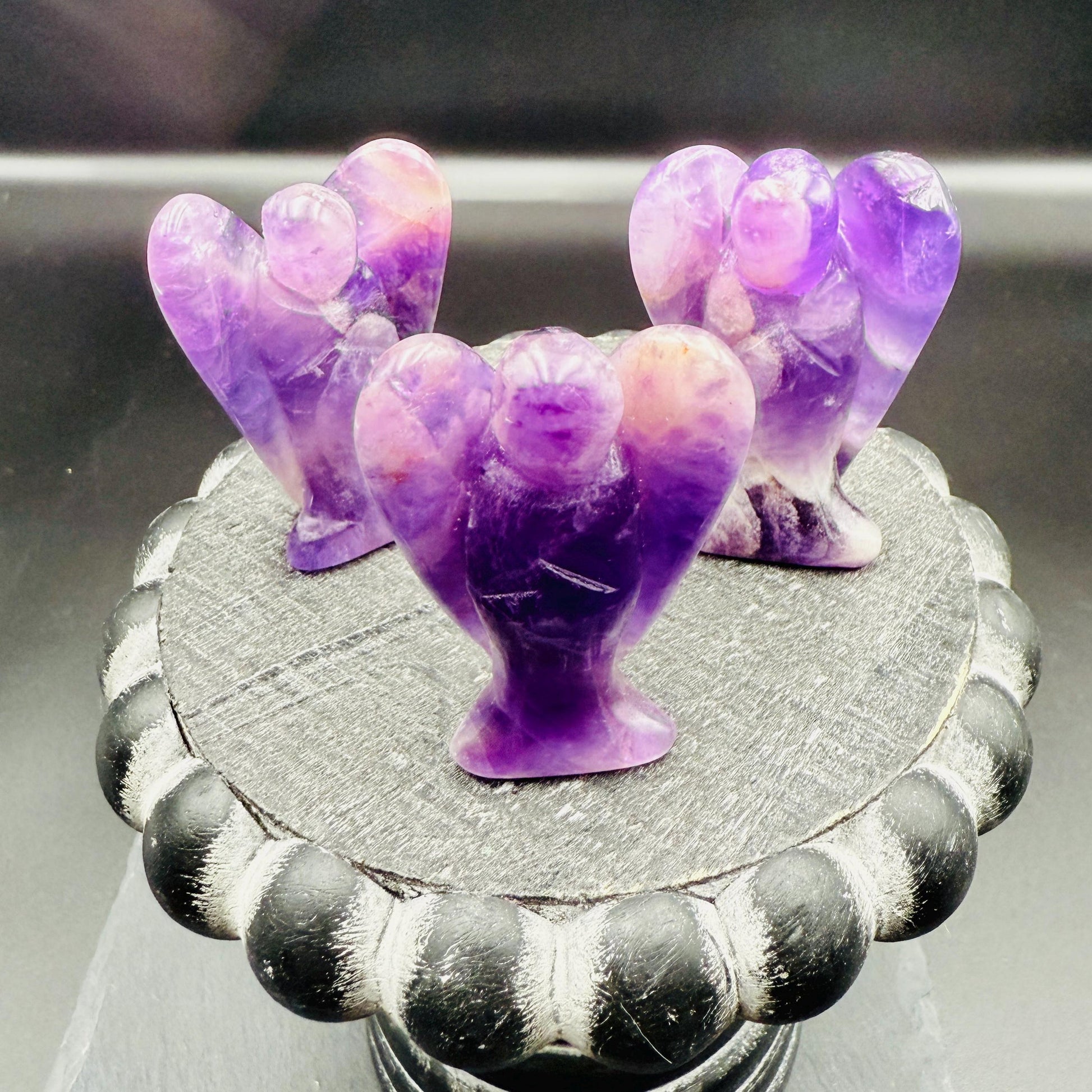 May include: Three amethyst angel figurines are displayed on a decorative, dark gray, round pedestal. The angels have purple and white coloring. The pedestal has a black and gray scalloped edge. The background is dark.