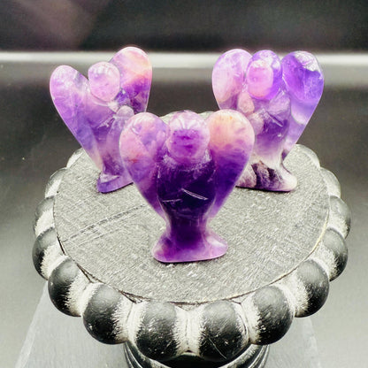 May include: Three amethyst angel figurines are displayed on a decorative, dark gray, round pedestal. The angels have purple and white coloring. The pedestal has a black and gray scalloped edge. The background is dark.