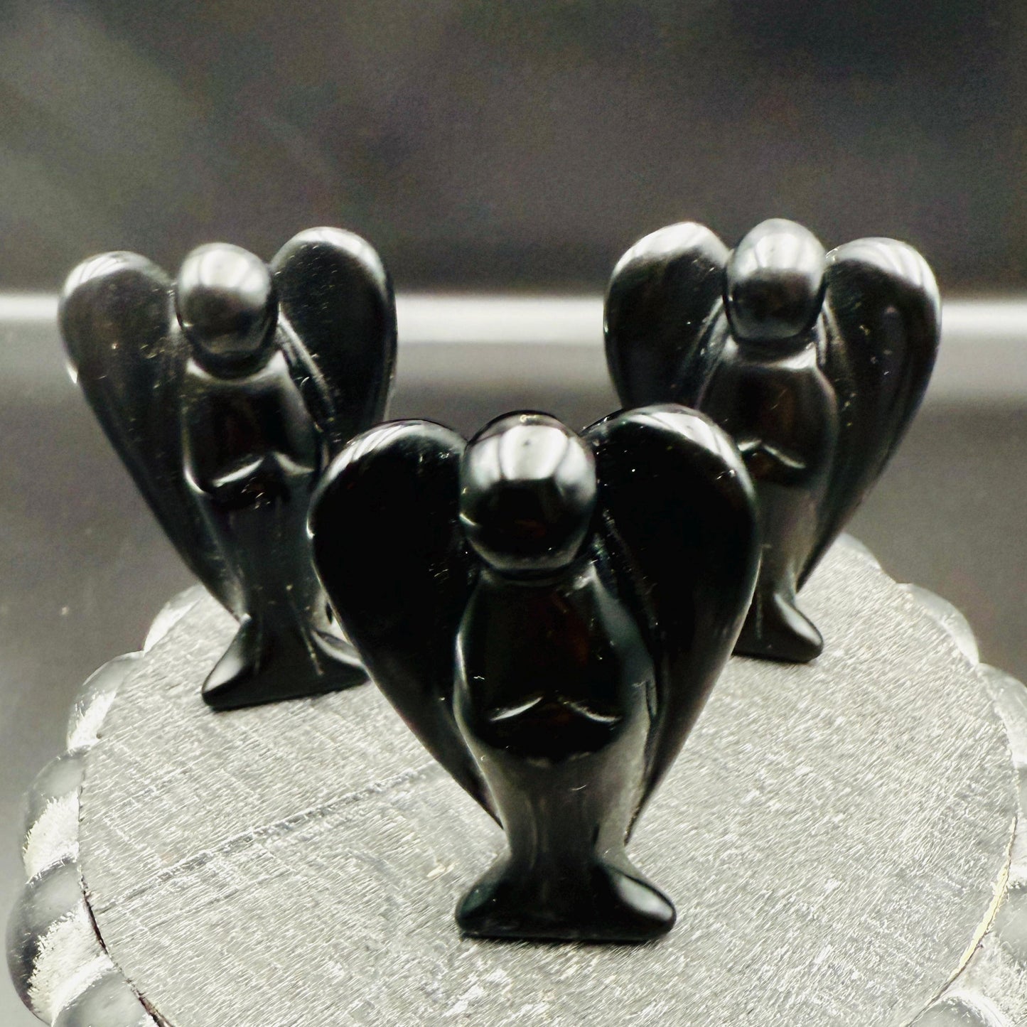May include: Three black obsidian angel figurines. Each angel has outstretched wings and is in a praying pose. The figurines are displayed on a textured, gray surface. The angels are a dark, glossy black.