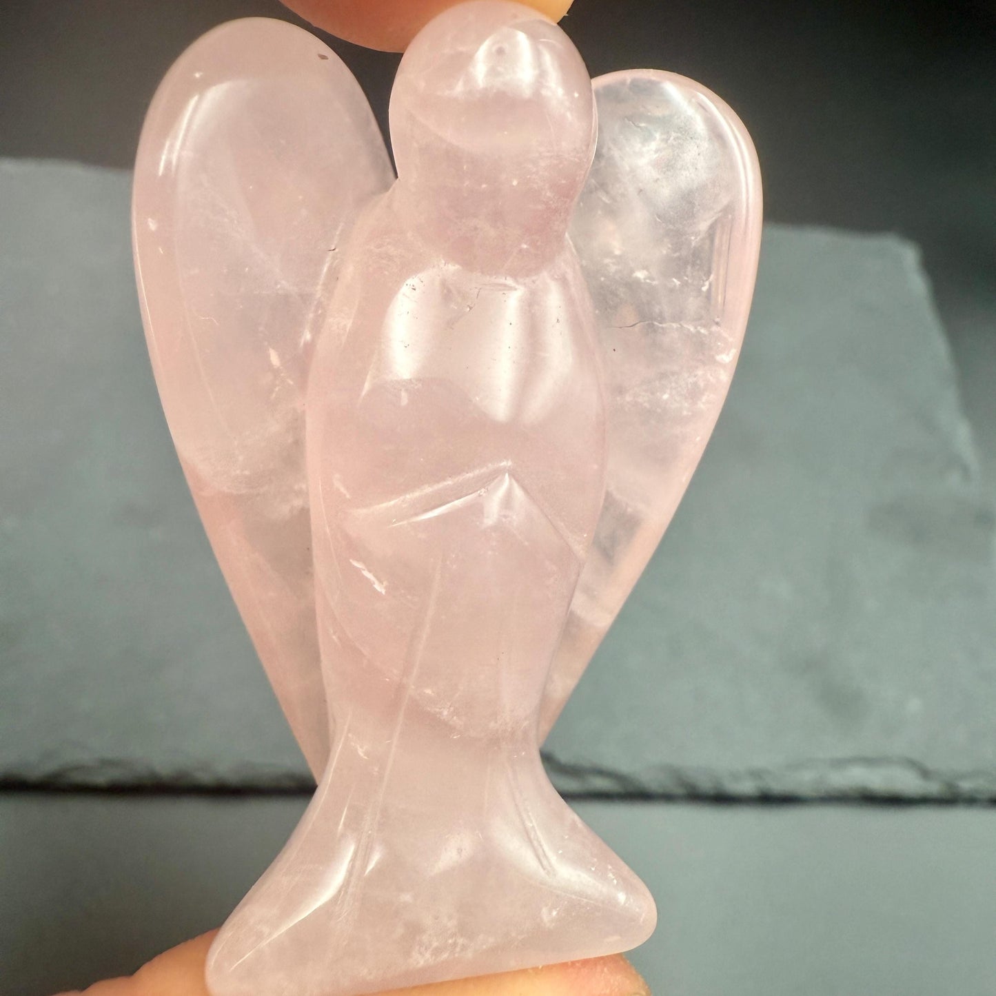 May include: A carved rose quartz angel figurine. The angel has outstretched wings and is wearing a long gown. The figurine is a soft pink color and is held by a person's fingers.