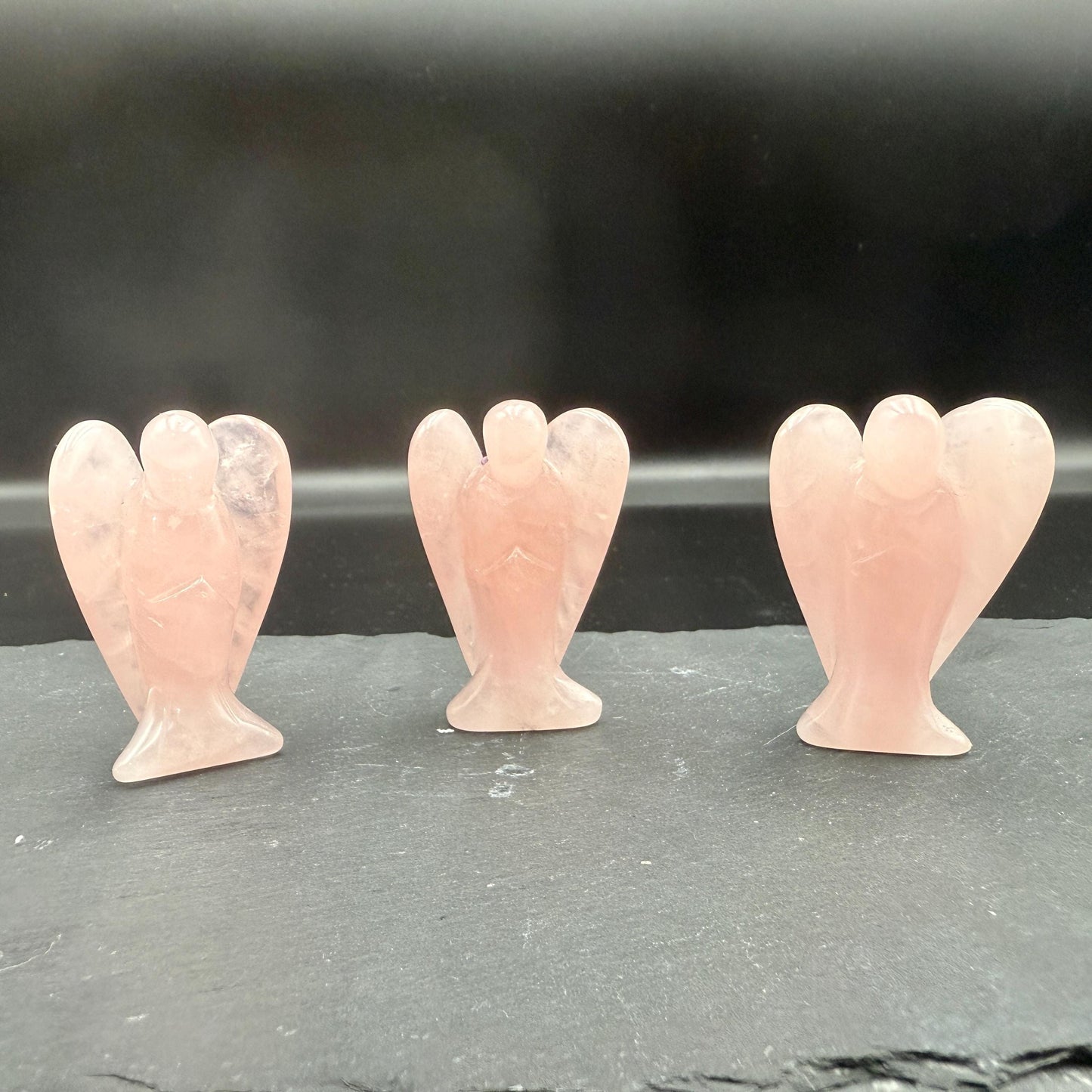 May include: Three rose quartz angel figurines. Each angel has outstretched wings and a flowing gown. The angels are a soft pink color and are displayed on a dark gray surface. The angels are a spiritual or decorative item.