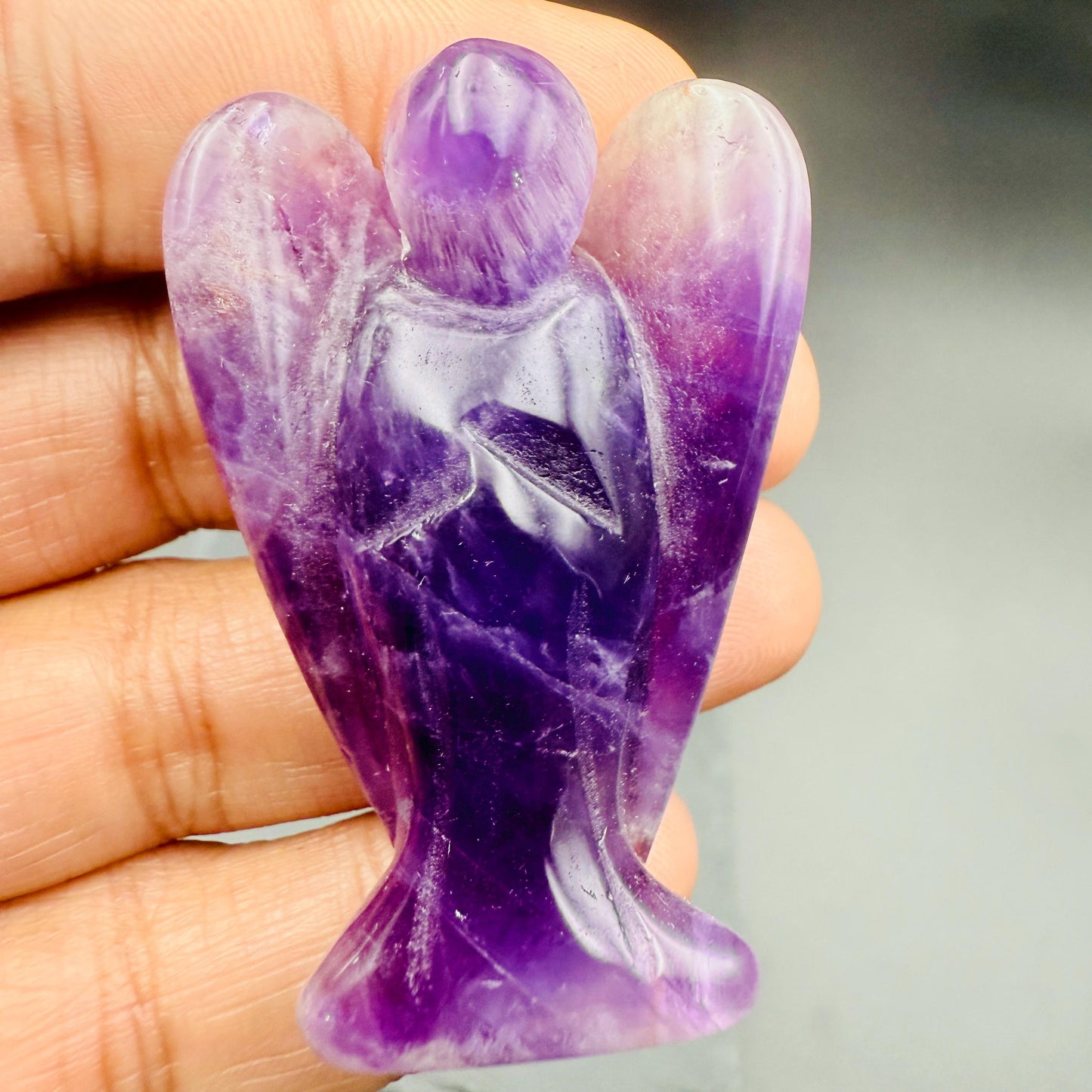 May include: A carved amethyst angel figurine. The angel has outstretched wings and is depicted in a standing pose. The stone is a deep purple color with lighter purple and white veining throughout. The figurine is approximately 3 inches tall.