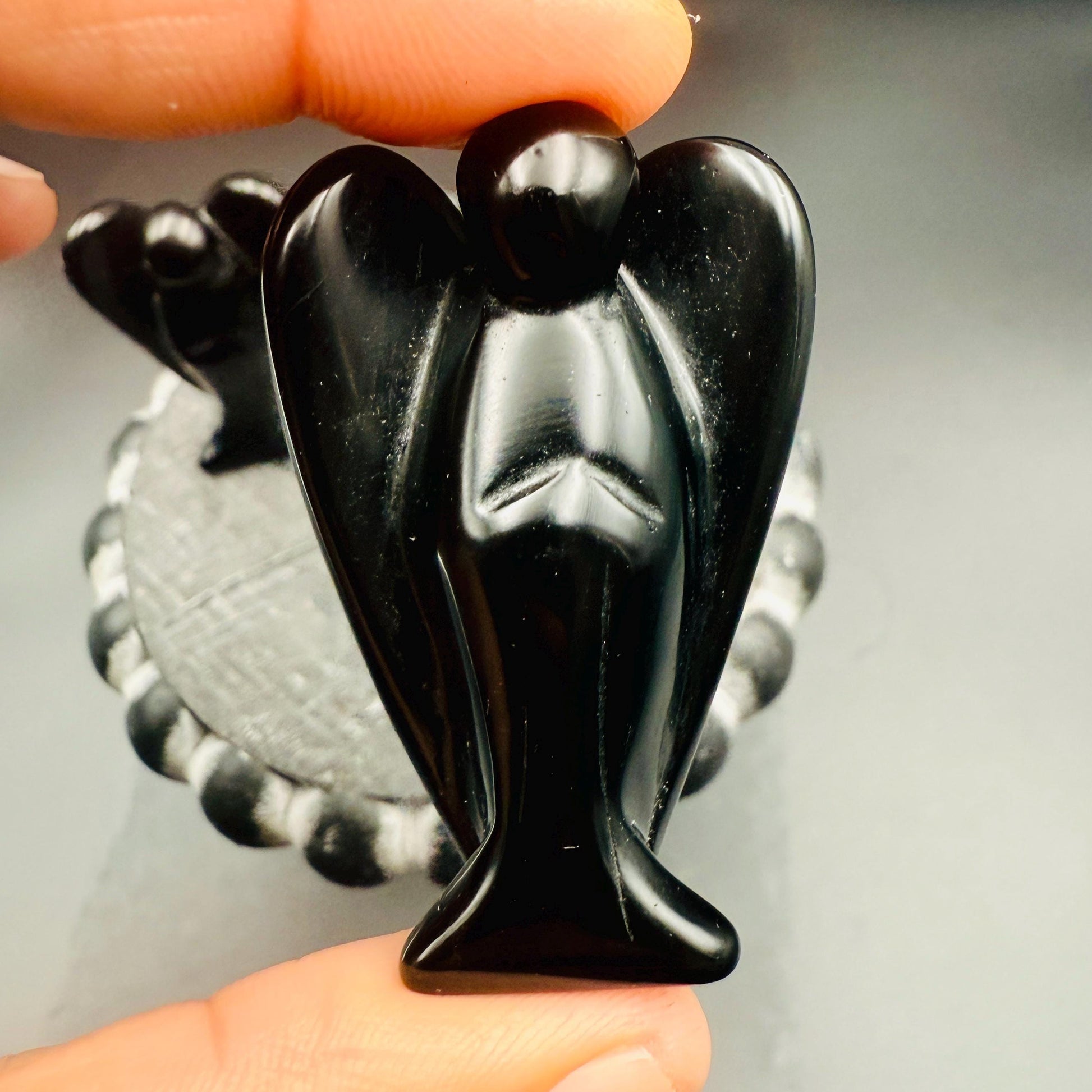 May include: A small, black angel figurine with outstretched wings. The angel is carved from a glossy, dark material and is held between fingers. The figurine has a smooth, polished surface and detailed wings.