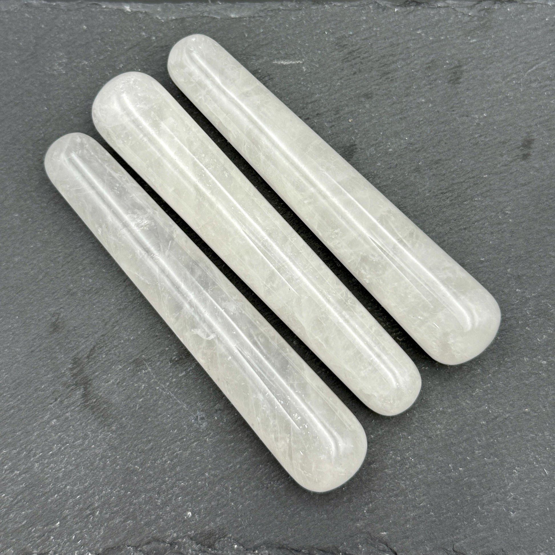 May include: Three translucent, cylindrical crystal wands are arranged diagonally on a dark gray surface. The wands have rounded ends and a smooth, polished texture. The crystals appear to be a pale, milky white color.