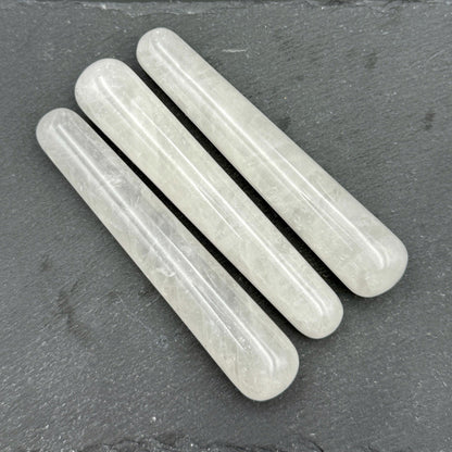 May include: Three translucent, cylindrical crystal wands are arranged diagonally on a dark gray surface. The wands have rounded ends and a smooth, polished texture. The crystals appear to be a pale, milky white color.
