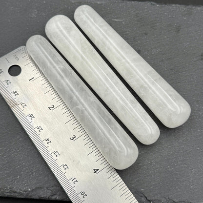 May include: Three light-colored, cylindrical stone massage tools are displayed diagonally. A metal ruler with inch markings is placed next to the tools. The tools appear to be approximately 4 inches long.