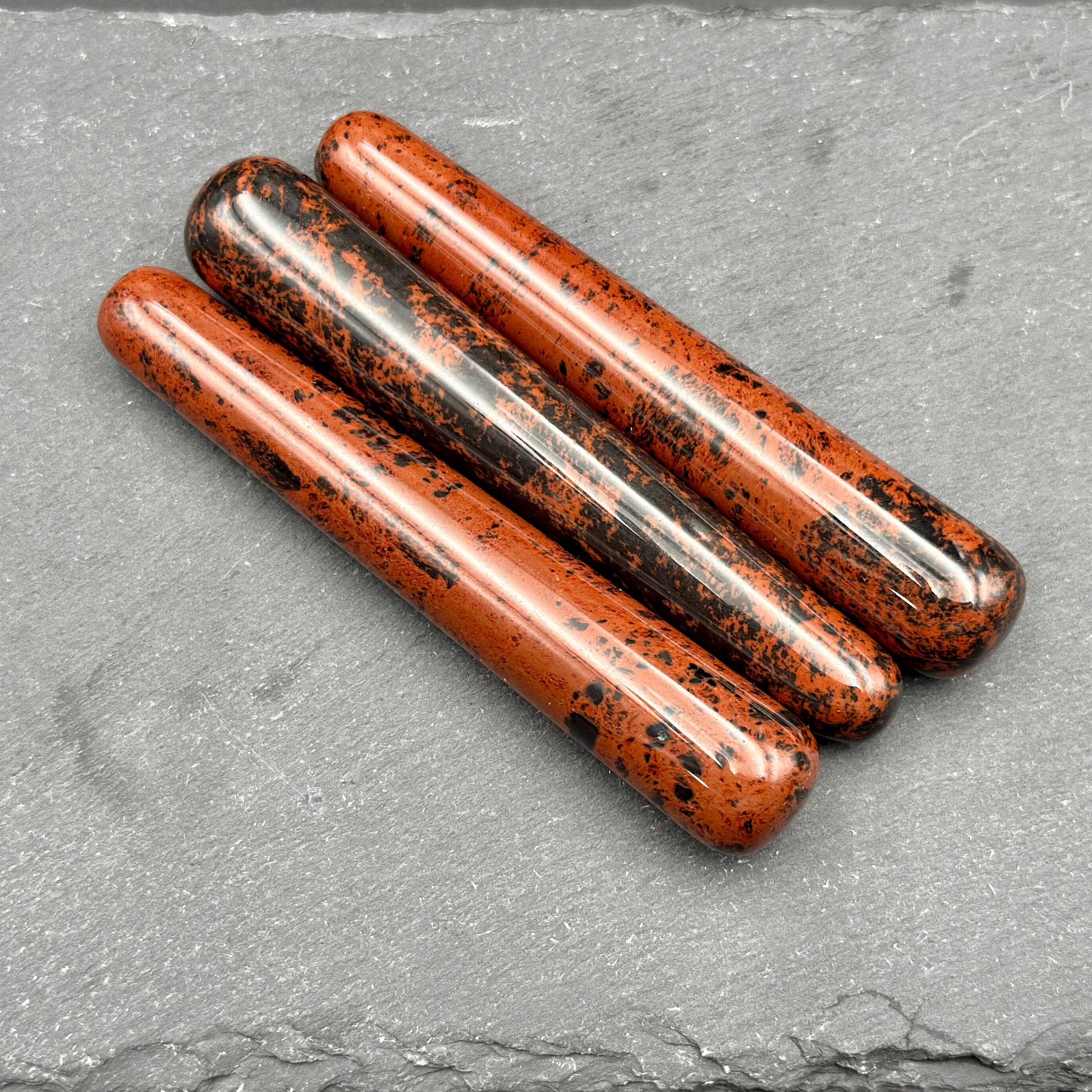 May include: Three polished obsidian wands with a reddish-brown base color and black speckles. The wands are cylindrical with rounded ends, lying diagonally on a dark gray surface. The wands are smooth and reflect light.