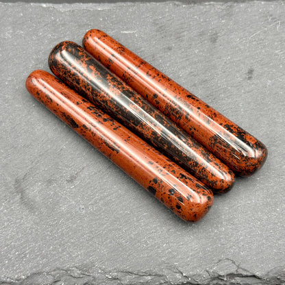May include: Three polished obsidian wands with a reddish-brown base color and black speckles. The wands are cylindrical with rounded ends, lying diagonally on a dark gray surface. The wands are smooth and reflect light.