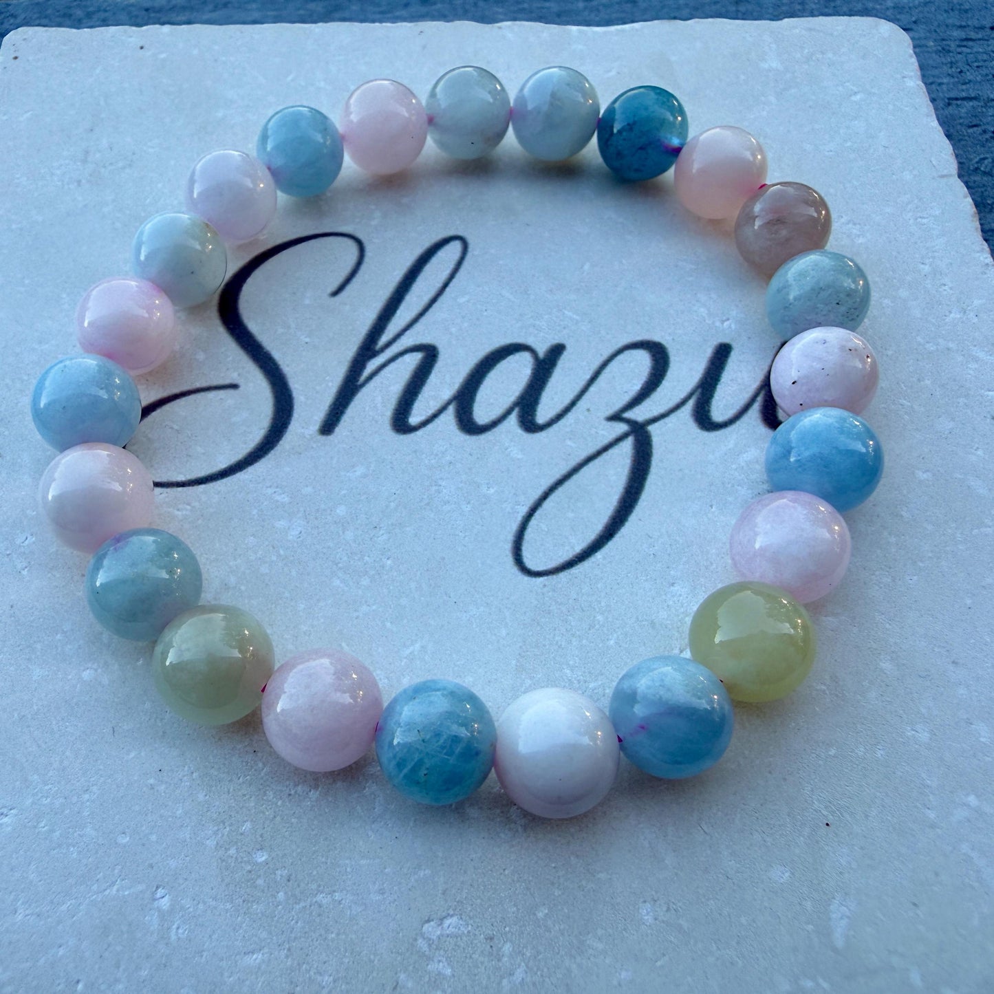 May include: A circular beaded bracelet featuring smooth, polished spherical beads in various pastel shades including light blue, soft pink, white, and pale yellow. The bracelet is arranged in a ring shape on a textured surface with the word Shazu printed.