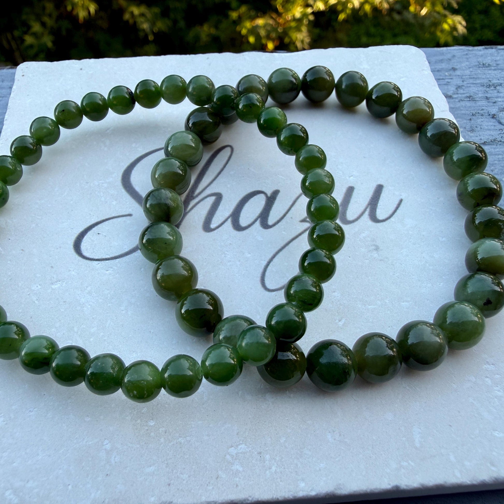 May include: Two circular bracelets composed of smooth, polished, dark green spherical beads. The beads are uniform in size and strung together on an elastic cord. The bracelets are arranged in an overlapping pattern on a white textured surface.