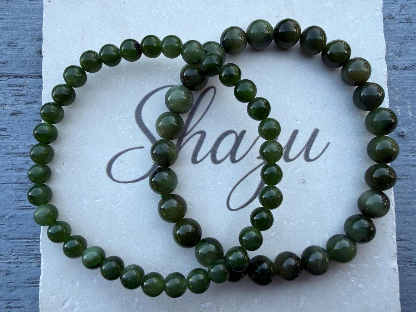 May include: Two circular bracelets made of polished, dark forest green spherical beads. The beads are uniform in size and strung together on an elastic cord. The bracelets are arranged in an overlapping pattern, creating a linked appearance.