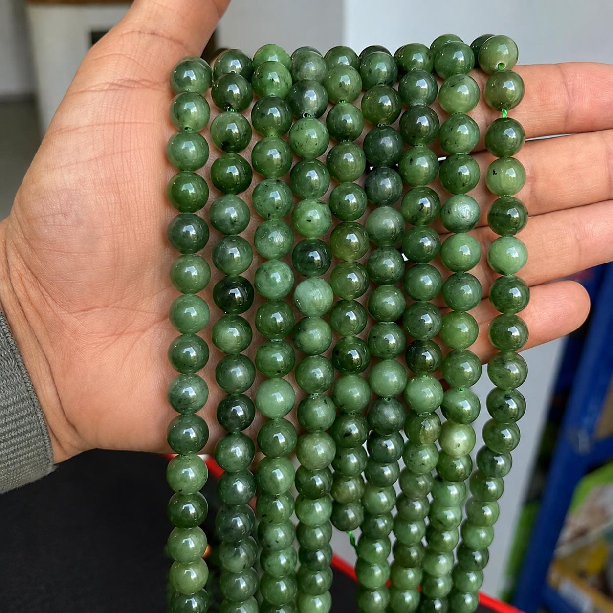 May include: A collection of smooth, round, dark green jade beads strung together on multiple vertical strands. Each bead features a polished, glossy finish with subtle natural variations in color, ranging from deep forest green to lighter olive tones.