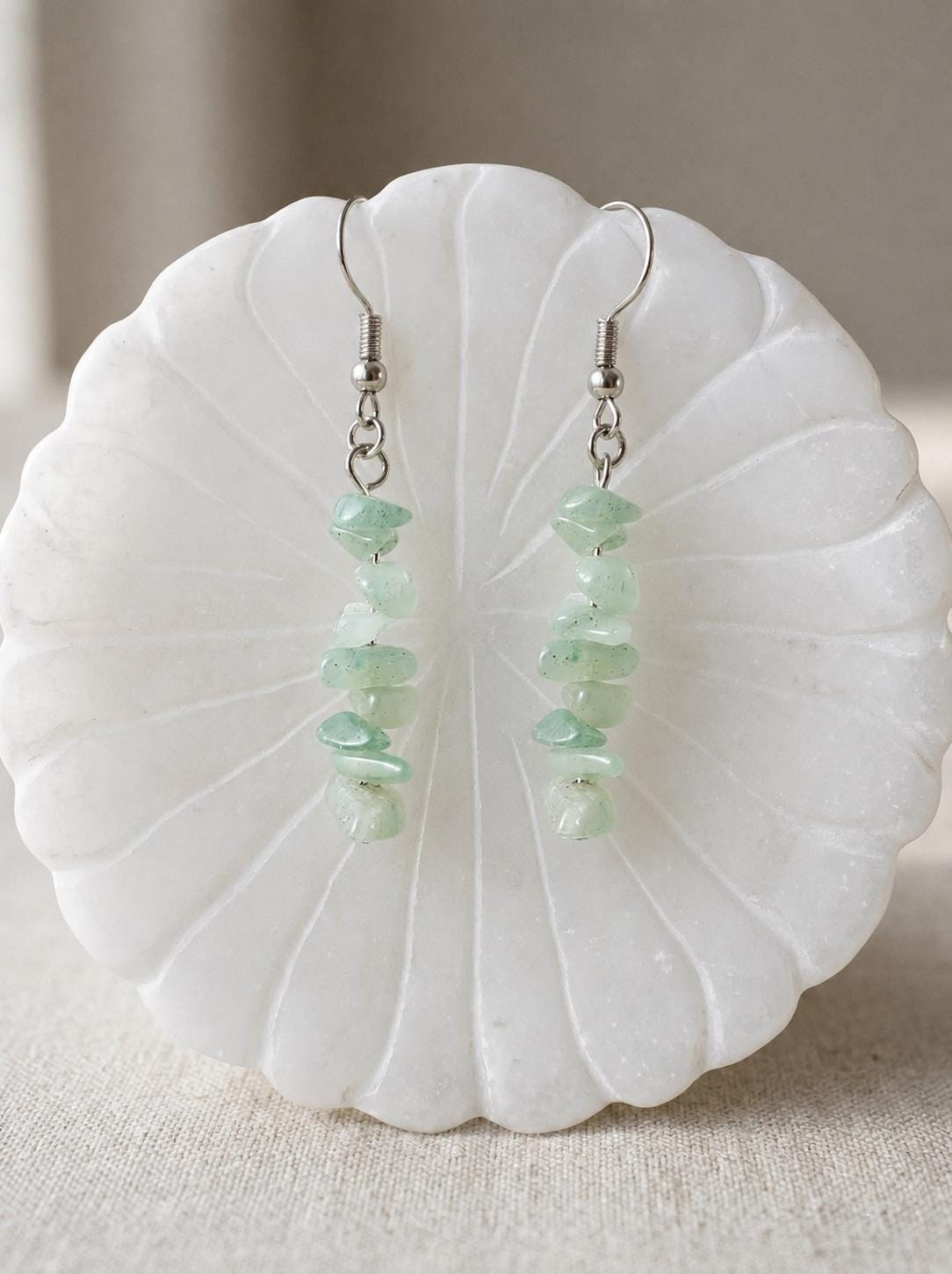 May include: A pair of dangle earrings featuring light green, irregular, tumbled stone chips strung vertically on silver-toned metal wire. Each earring is finished with a simple silver-toned hook finding for pierced ears, showcasing a natural texture.
