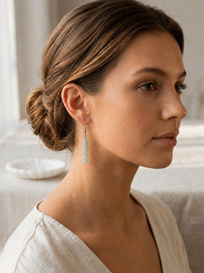May include: A drop earring featuring a vertical arrangement of small, pale green, tumbled stone chips suspended from a silver-toned metal hook. The earring hangs straight down, showcasing the light green color and irregular texture of the stones.