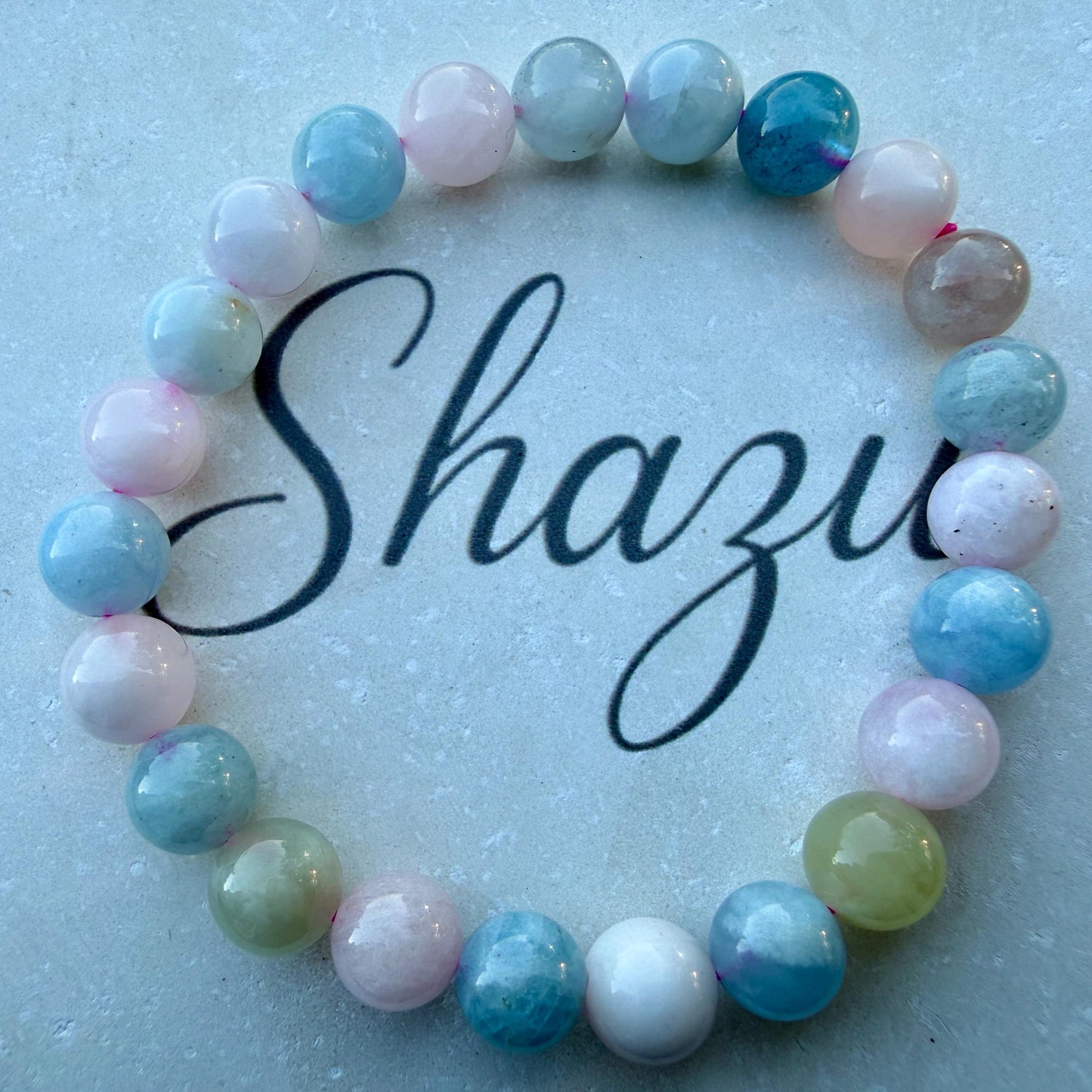 May include: A circular beaded bracelet featuring smooth, polished round beads in shades of light blue, soft pink, white, and pale green. The beads are strung together in a repeating pattern, and the word Shazu is written in black script in the center.