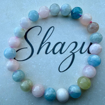 May include: A circular beaded bracelet featuring smooth, polished round beads in shades of light blue, soft pink, white, and pale green. The beads are strung together in a repeating pattern, and the word Shazu is written in black script in the center.