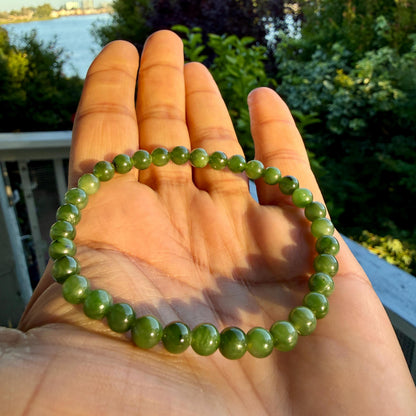 May include: A circular bracelet composed of small, smooth, spherical green beads. The beads exhibit varying shades of translucent and opaque olive green, strung together in a continuous loop to form a simple, uniform band of polished stone.
