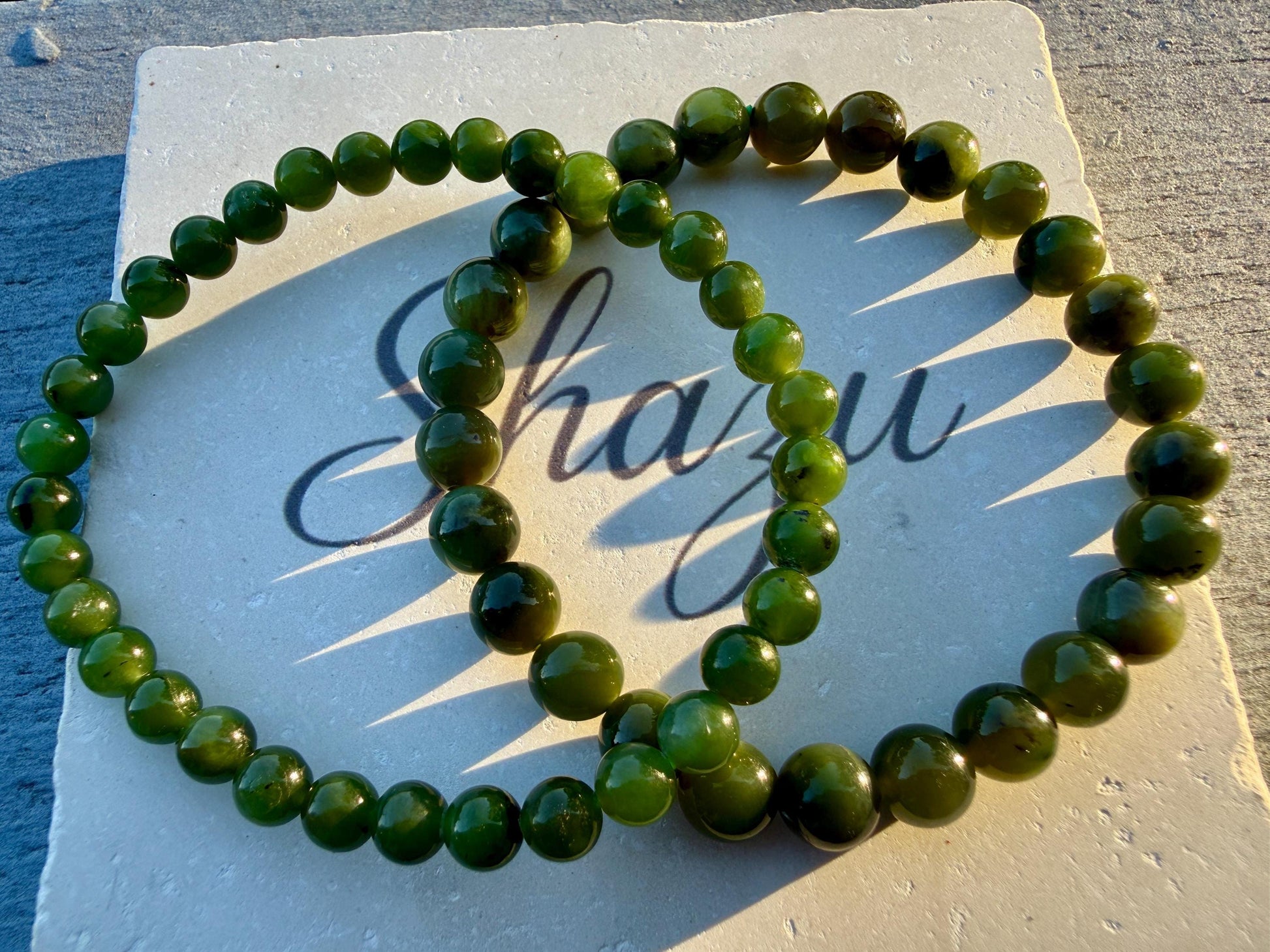 May include: Two circular bracelets composed of smooth, polished, dark green spherical beads. The bracelets are arranged in an overlapping pattern on a square, off-white stone tile that features the cursive text Shazu printed in black ink.