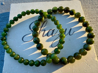 May include: Two circular bracelets composed of smooth, polished, dark green spherical beads. The bracelets are arranged in an overlapping pattern on a square, off-white stone tile that features the cursive text Shazu printed in black ink.