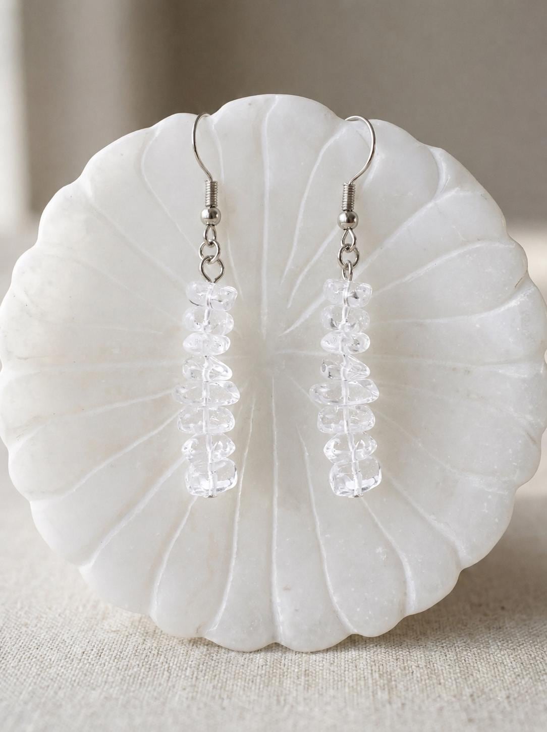 May include: A pair of dangle earrings featuring silver-toned metal hooks and vertical strands of small, clear, irregular-shaped crystal beads. The earrings hang straight down, showcasing the translucent, faceted texture of the clear stones.