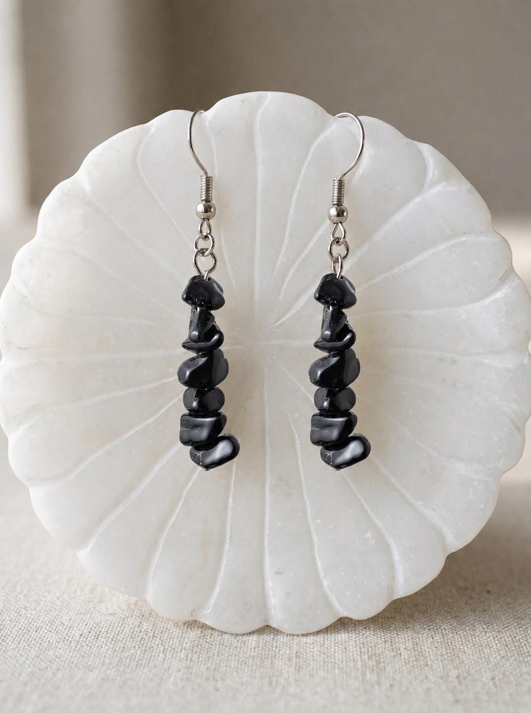 May include: A pair of dangle earrings featuring vertical strands of polished black stone chips suspended from silver-toned metal fishhook wires. Each earring consists of multiple irregular, smooth black stone pieces stacked to create a linear, textured drop design.