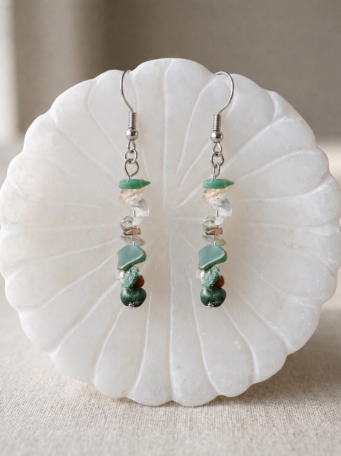 May include: A pair of dangle earrings featuring silver-toned metal hooks and a vertical arrangement of small, irregular, tumbled gemstone chips in shades of green, clear, and brown, suspended in a linear design from the top metal loop.