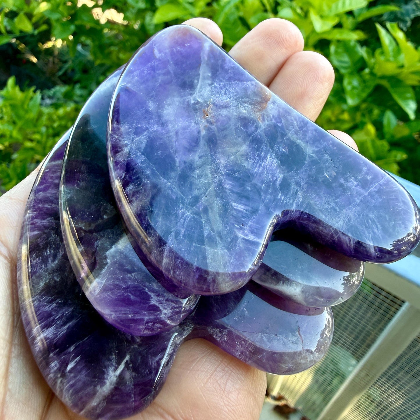 May include: A stack of three polished, heart-shaped amethyst stones. Each stone features a deep purple hue with natural white crystalline inclusions and a smooth, glossy surface. The stones are layered, showing variations in color intensity.