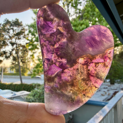 May include: A heart-shaped polished stone featuring translucent layers of deep purple, violet, and golden-brown mineral inclusions. The surface is smooth and glossy, allowing light to pass through the crystalline structure to reveal internal patterns.