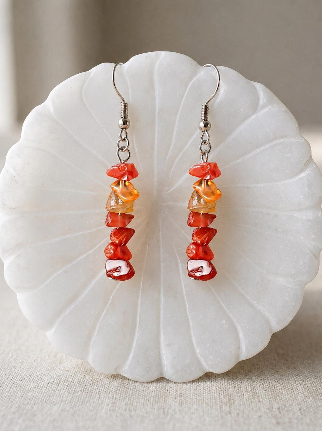 May include: A pair of dangle earrings featuring a vertical arrangement of small, irregular, polished stone chips in shades of vibrant orange, deep red, and translucent amber, suspended from silver-toned metal fishhook ear wires.