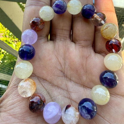 May include: A stretch bracelet composed of smooth, round, multi-colored gemstone beads. The beads feature a variety of translucent and opaque finishes in shades of deep purple, pale lavender, golden yellow, clear white, and dark reddish-brown.