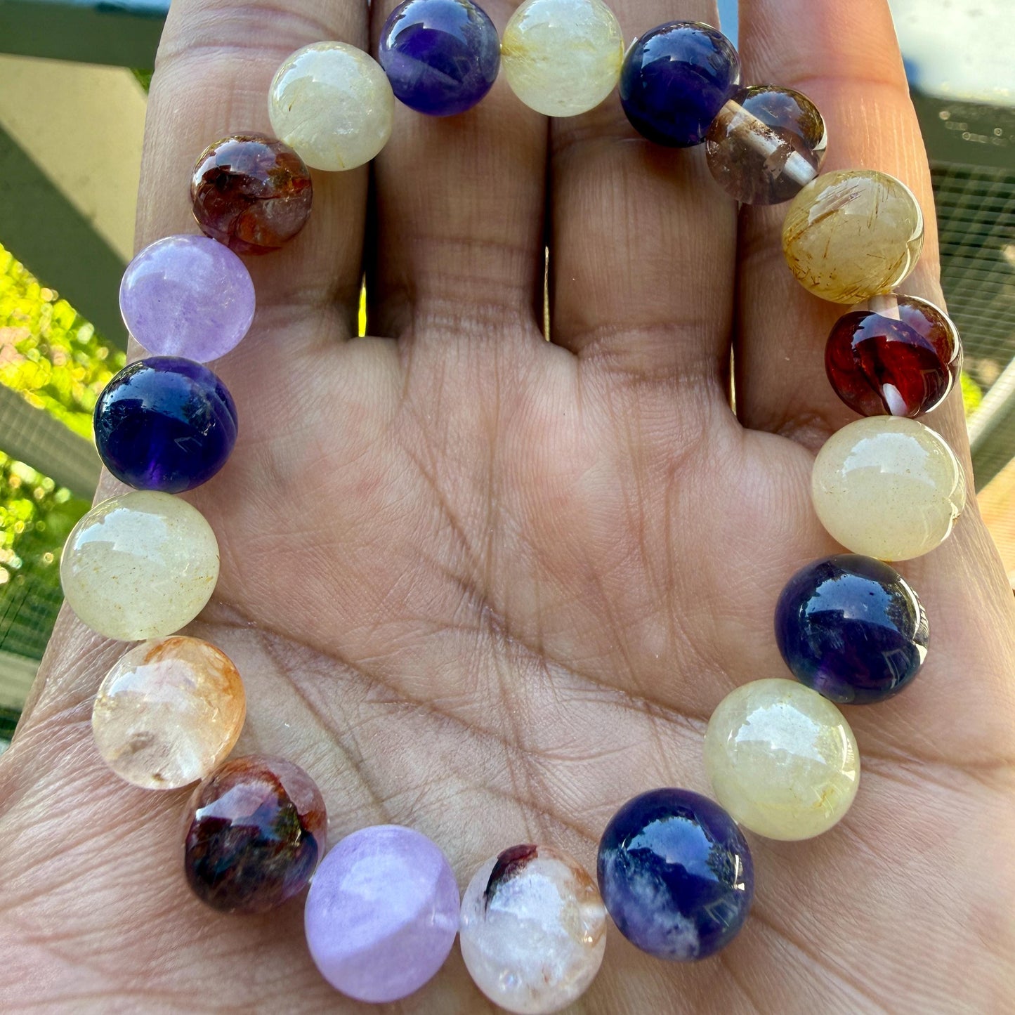 May include: A beaded bracelet featuring smooth, round gemstone beads in a variety of colors including deep purple, translucent white, golden yellow, and dark reddish-brown. The beads are arranged in a circular pattern, showcasing natural variations.
