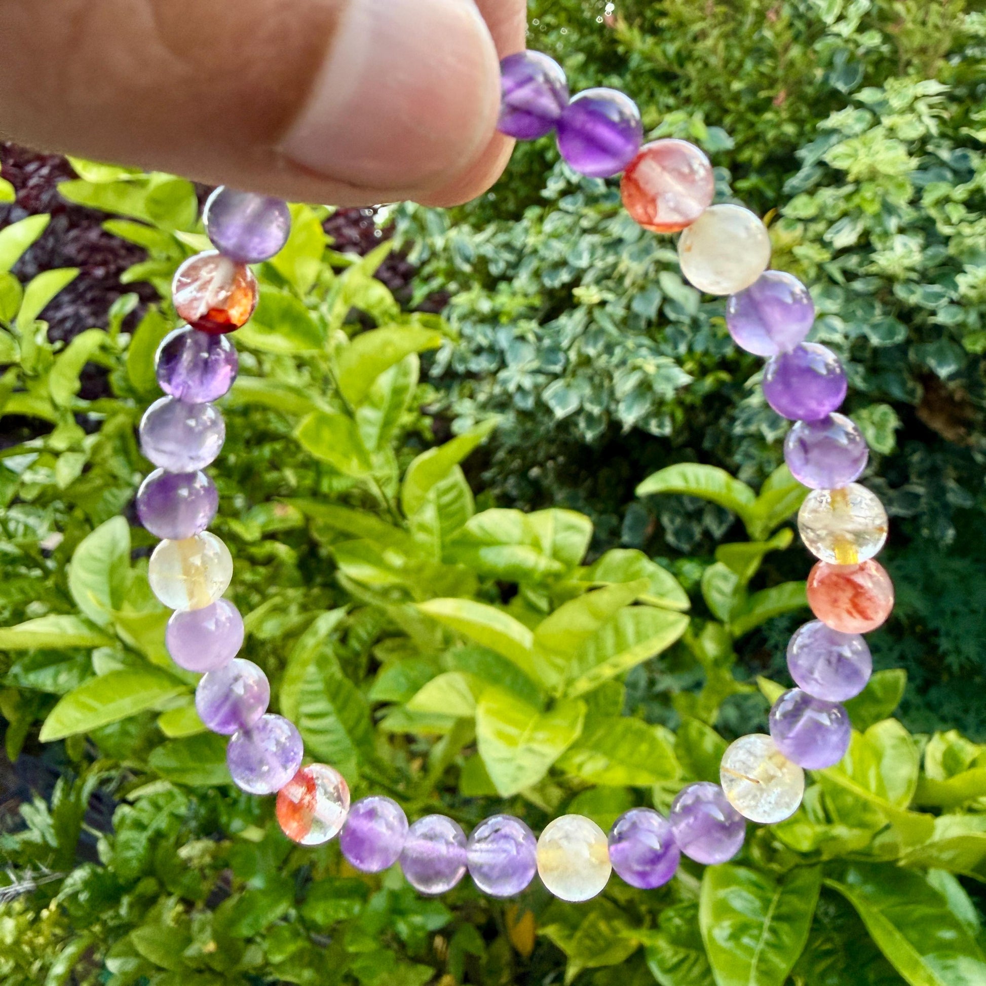 May include: A beaded bracelet featuring round polished stones in shades of purple, translucent yellow, and orange-red. The beads are strung together in a circular pattern, showcasing variations in color intensity and natural stone clarity throughout.