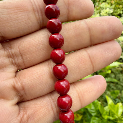 May include: A vertical strand of smooth, round, glossy red beads. Each bead is uniform in size and shape, featuring a vibrant, solid red color with a reflective surface finish. The beads are strung together in a single, straight line, showcasing a consistent, polished appearance throughout the entire length of the visible beaded strand.