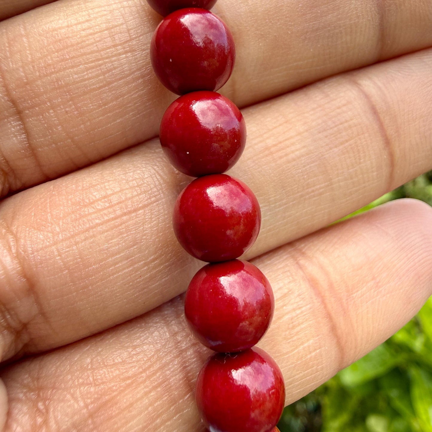 May include: A strand of smooth, spherical red beads arranged in a vertical line. Each bead features a glossy, polished finish that reflects light, creating bright highlights on the surface of the deep red spheres. The beads are uniform in size.