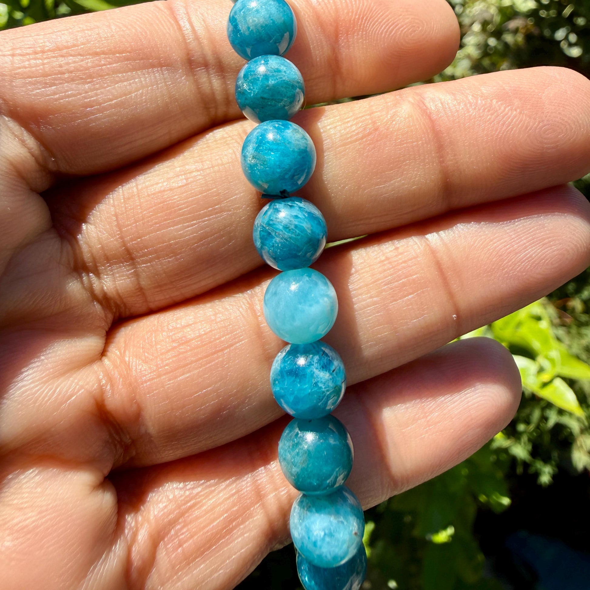 May include: A strand of round, polished blue apatite beads. The beads exhibit varying shades of vibrant teal and sky blue, featuring natural internal inclusions and a smooth, glossy finish that reflects light across their spherical surfaces.