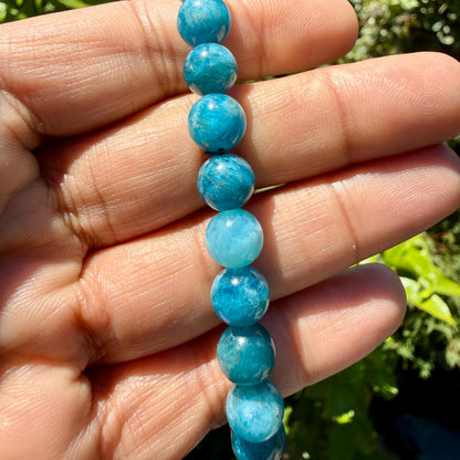 May include: A strand of round, polished blue apatite beads. The beads exhibit varying shades of vibrant teal and sky blue, featuring natural internal inclusions and a smooth, glossy finish that reflects light across their spherical surfaces.