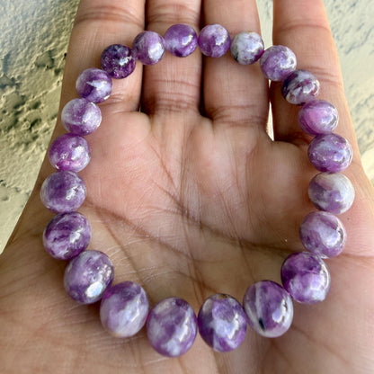 May include: A stretch bracelet composed of round, polished beads in varying shades of purple, lavender, and white. The beads feature natural marbling and swirling patterns throughout, creating a smooth, glossy texture across the entire circular piece.
