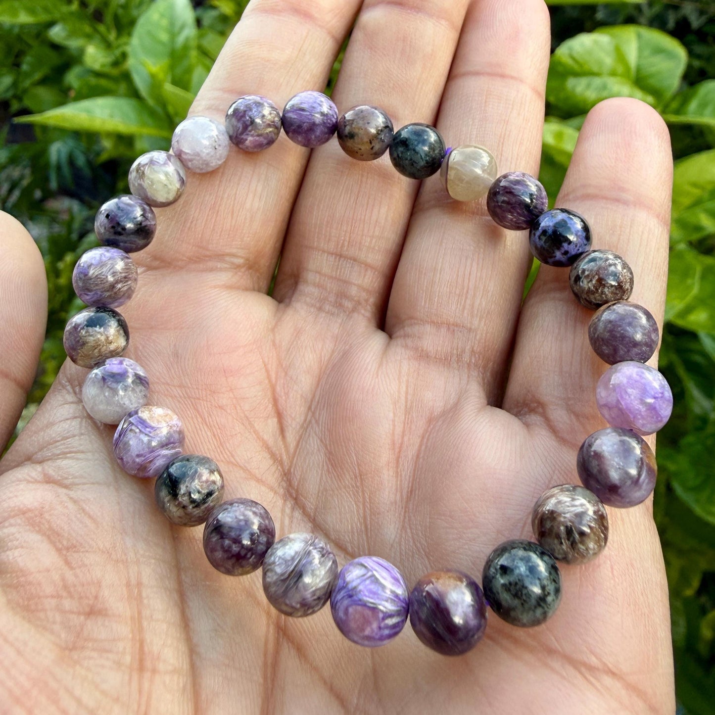 May include: A stretch bracelet composed of round, polished beads in varying shades of purple, lavender, dark grey, and translucent white. The beads feature natural swirling patterns and inclusions, strung together on a flexible cord to form a circular loop.