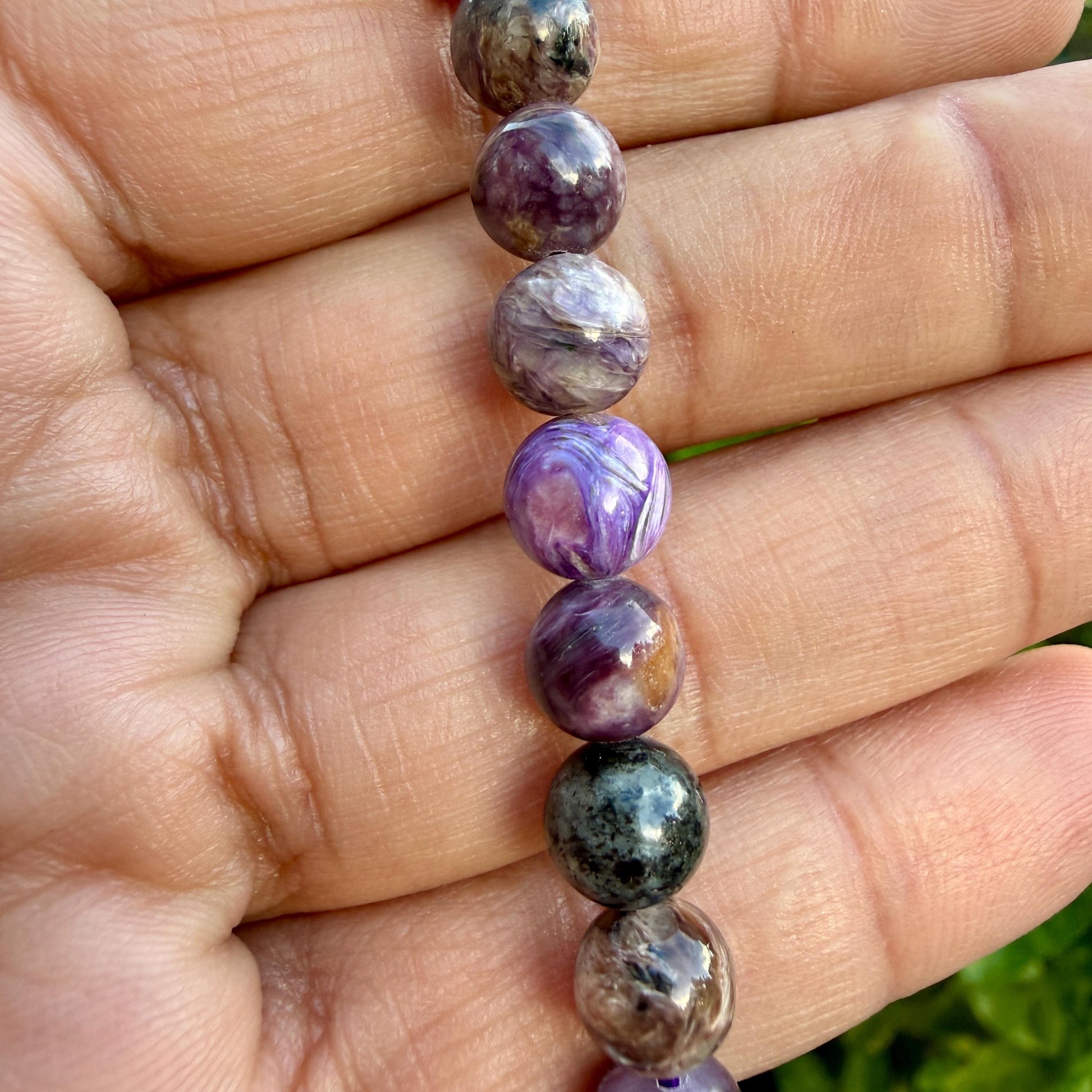 May include: A strand of spherical beads featuring a variety of natural stone patterns in shades of deep purple, lavender, charcoal gray, and white. The beads exhibit swirling, marbled textures and polished surfaces arranged in a vertical linear sequence.