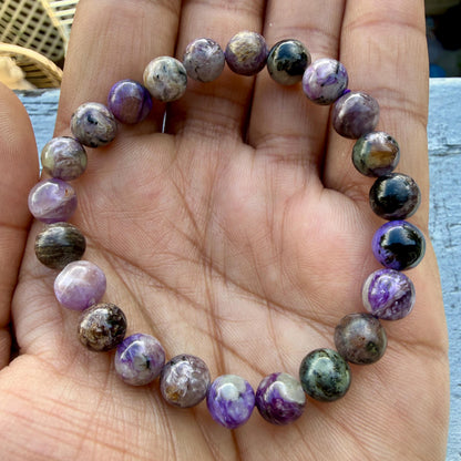 May include: A beaded bracelet featuring round, polished stones in shades of purple, lavender, black, and grey. The beads exhibit natural swirling patterns and varying levels of opacity, strung together in a circular loop to form a continuous band.