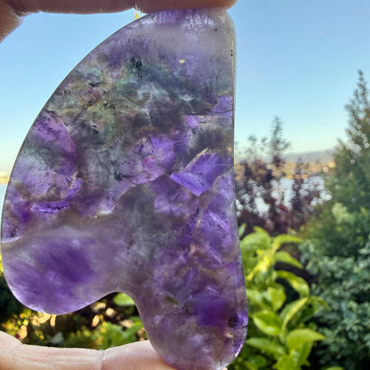 May include: A polished, translucent stone tool with a curved, asymmetrical shape. The surface displays a mix of deep purple, violet, and clear crystalline sections with internal fractures and natural color variations throughout the material.
