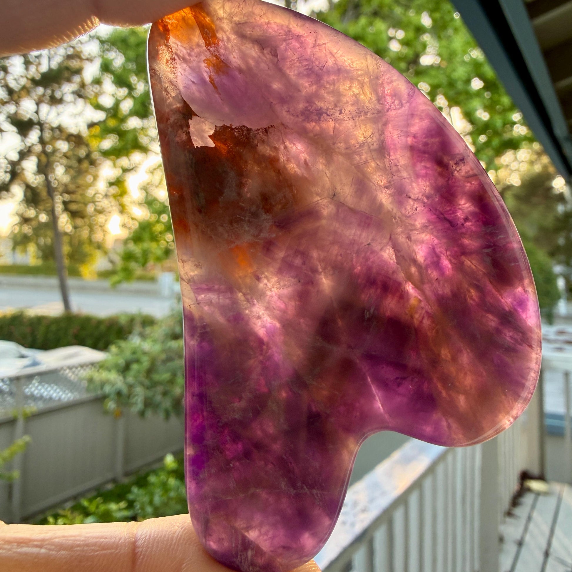 May include: A heart-shaped gua sha tool crafted from translucent purple and orange fluorite stone. The polished surface displays natural mineral inclusions, color gradients ranging from deep violet to amber, and a smooth, curved edge profile.